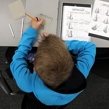 overhead shot of a boy sitting at a desk drawing human noses
